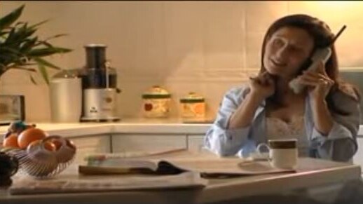 A woman sits at a kitchen table holding a cordless phone