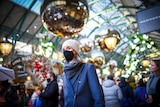 A young woman in a blue coat, black face mask and cream beanie walks through a crowded market