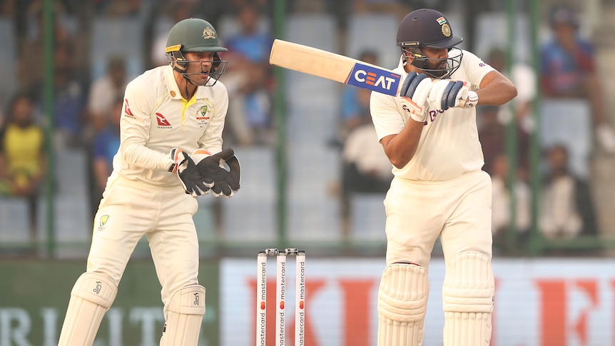 An Indian male Test batter hits a shot towards the leg side as the Australian wicketkeeper looks on.