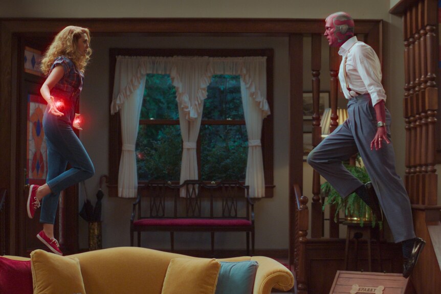 A 30-something woman and a masked man wearing 80s-style clothes are hovering in the air, looking at each other.