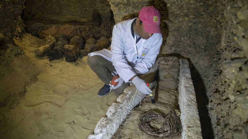 An archaeologist wearing a red cap uses a brush to remove dirt from a mummy.