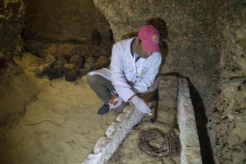 An archaeologist wearing a red cap uses a brush to remove dirt from a mummy.