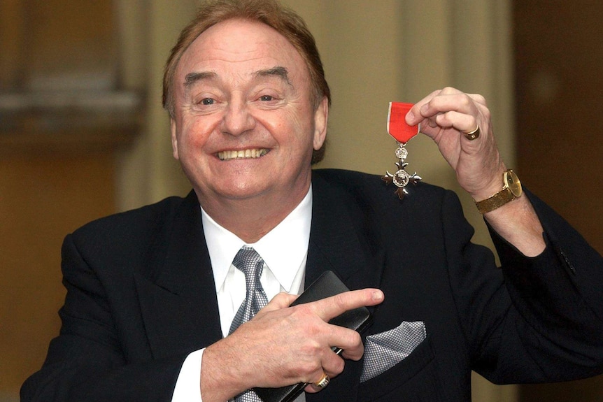 In this December 12, 2003 file photo, Gerry Marsden holds his MBE. He is smiling wearing a suit, pointing at the medal.