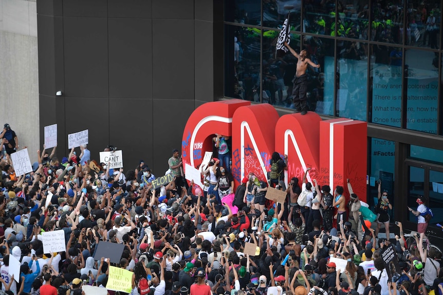 The red CNN logo is seen outside the broadcaster's building, with some people on it and hundreds standing in front of it.