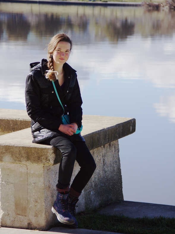 A teenaged girl sits beside a lake smiling, her hair in a braid.