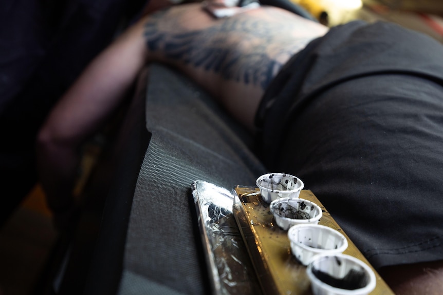 Bird's eye view of four black ink pots next to a man's hip, which is heavily tattooed. 