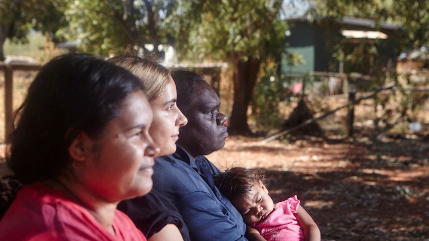 Three women sit in a line, one of them holding a baby, gazing into the distance.