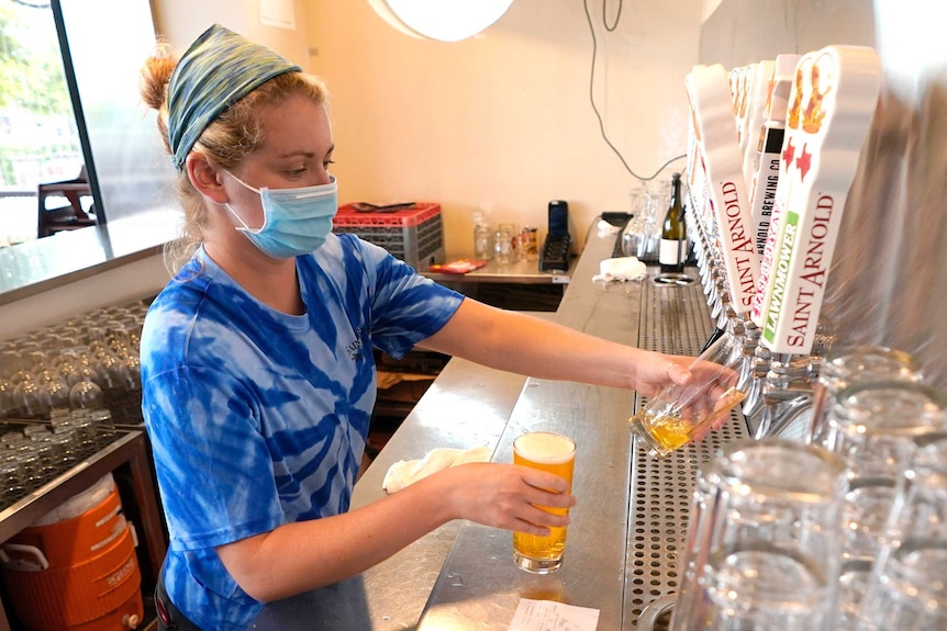 A bartender pours a beer from a tap while wearing a blue face mask.