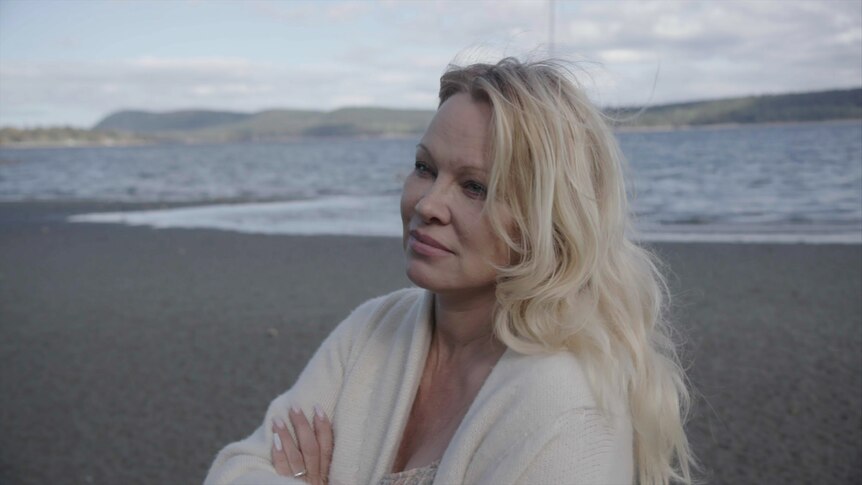 A side view of Pamela Anderson on a beach in a sweater looking solemn