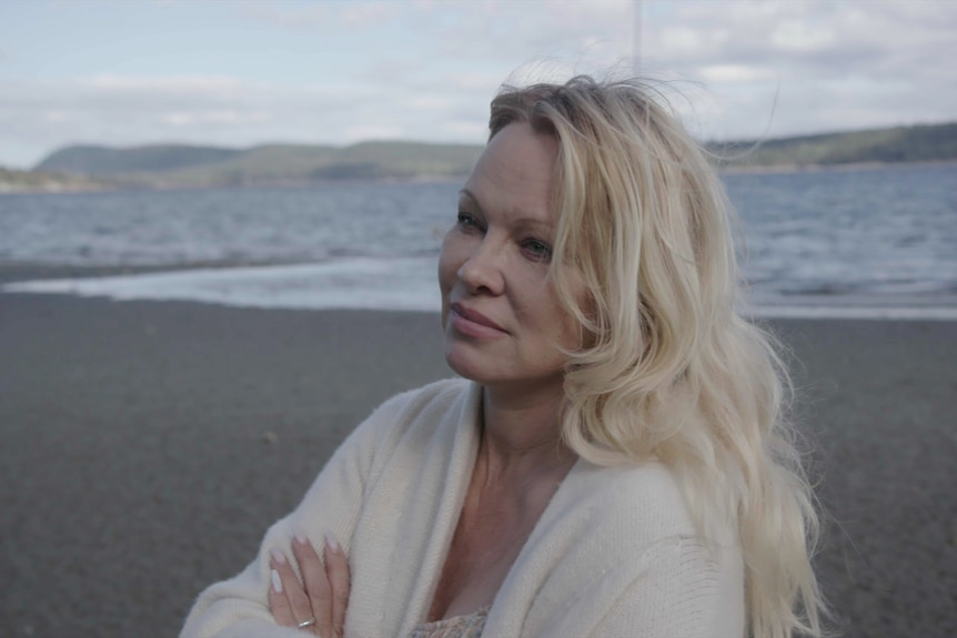 A side view of Pamela Anderson on a beach in a sweater looking solemn