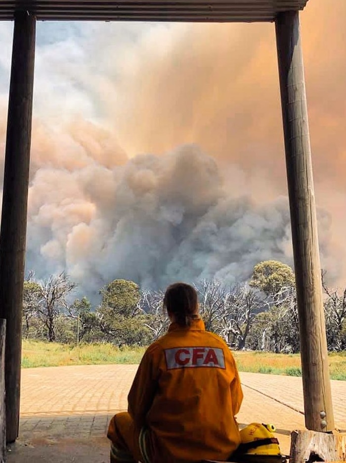A woman in a CFA yellow uniform shown before behind as she watches a giant cloud of bushfire smoke.