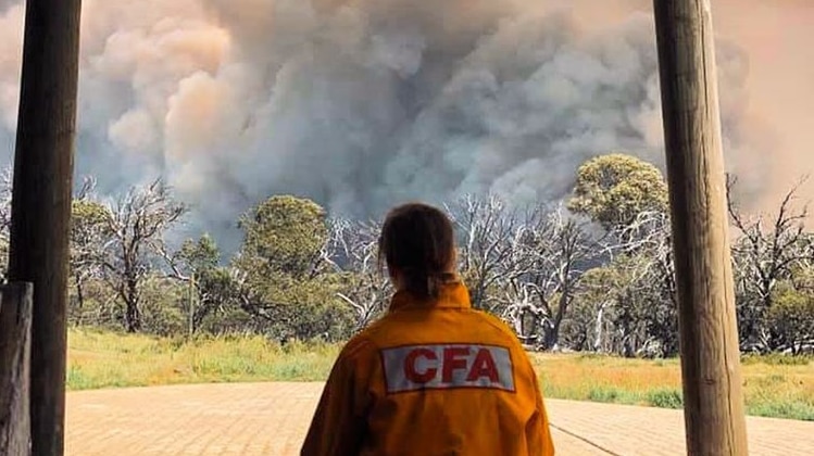 A woman in a CFA yellow uniform shown before behind as she watches a giant cloud of bushfire smoke.
