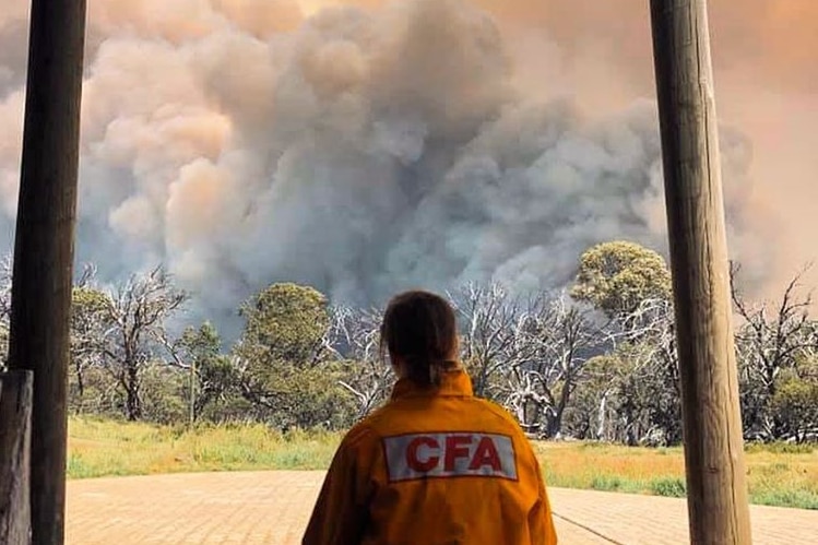 A woman in a CFA yellow uniform shown before behind as she watches a giant cloud of bushfire smoke.