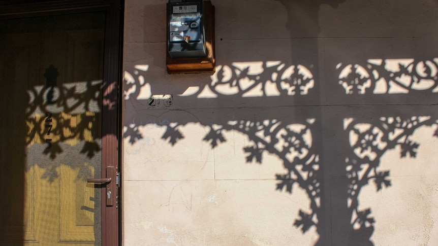 The shadow of iron lace on the wall beige of a Fitzroy terrace house, the yellow front door and an electrical box.