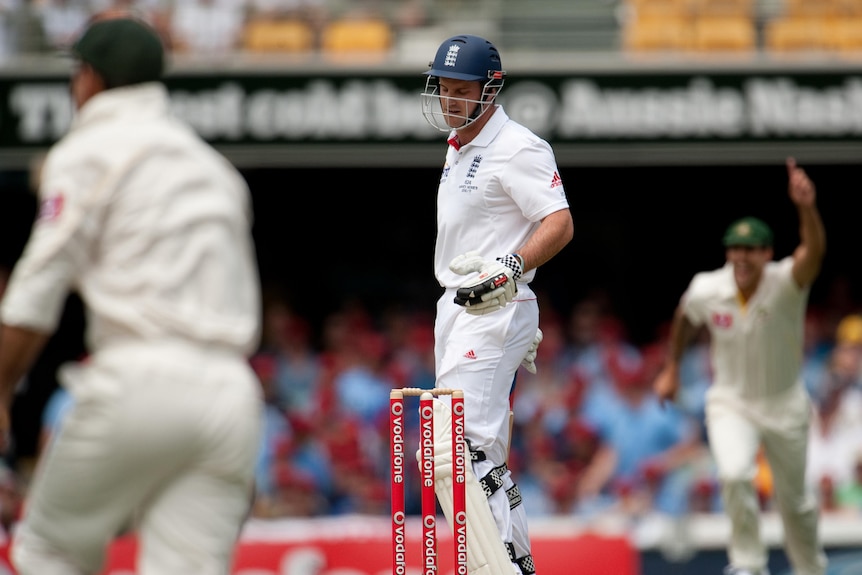 A dejected England captain looks down after being dismissed on day one of the Ashes as Australians celebrate in the background.