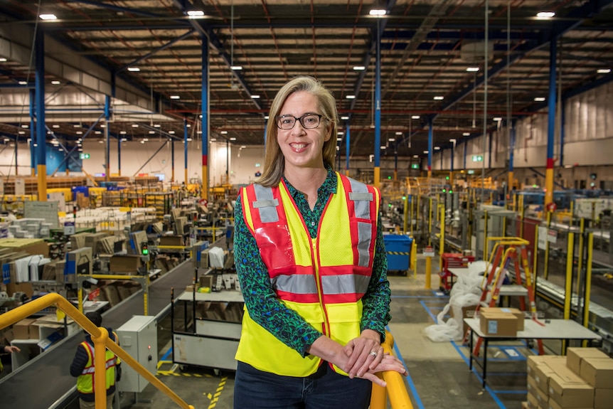 A woman with blonde hair and glasses and wearing a hi vis vest stands at the top of a set of yellow stairs in the Amazon wareho