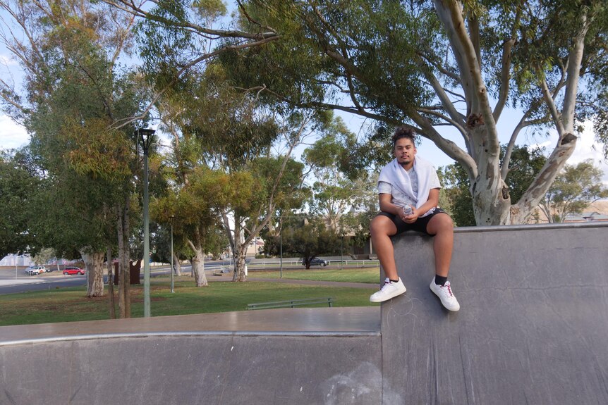 A young man sits on a skate park in front of some trees