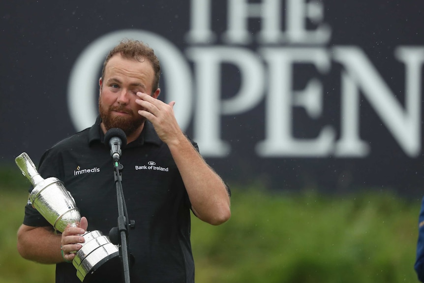 Shane Lowry, standing at a microphone, wipes at his eye with his left hand while holding a trophy with his right.