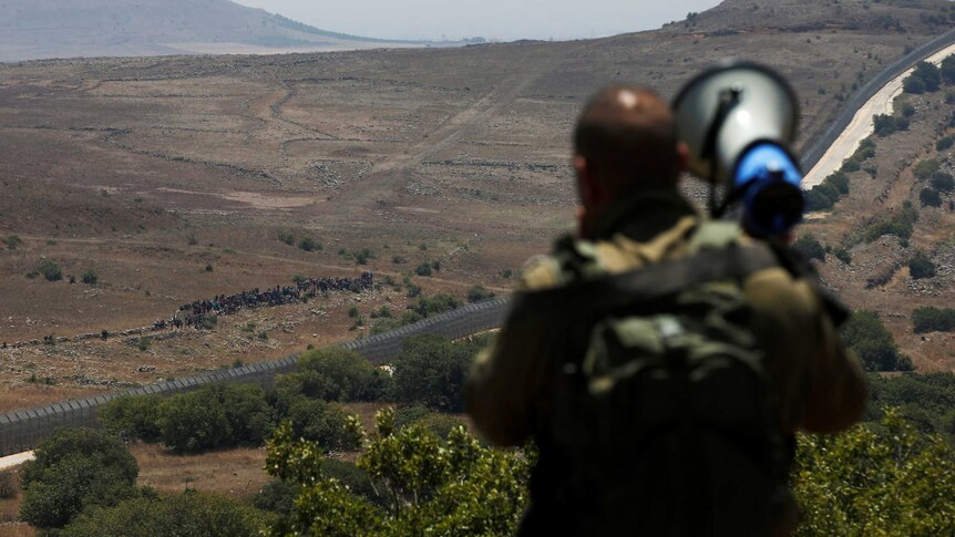 An Israeli soldier with a megaphone is in the foreground and a group of people are seen in the distance near a border fence.