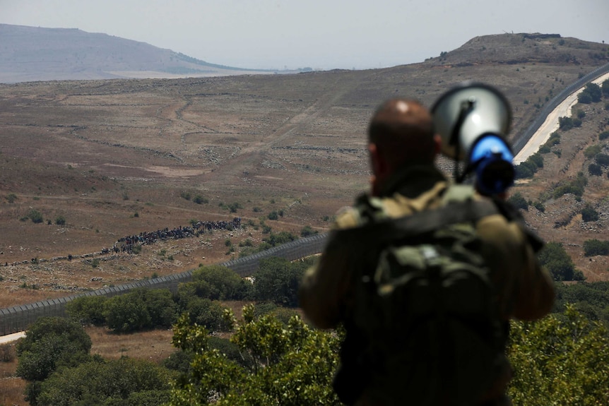 An Israeli soldier with a megaphone is in the foreground and a group of people are seen in the distance near a border fence.