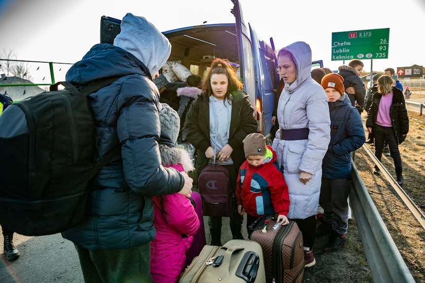 A group of young children in big coats stand next to a van on a roadside 