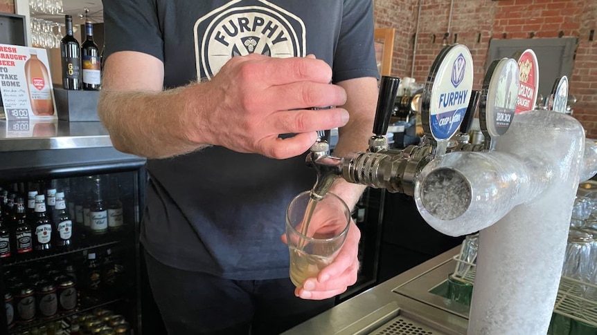 A man pours a Furphy beer from an icy tap behind the bar at a country pub.