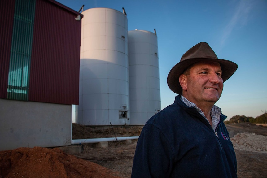 A man in an Akubra-style hat stands in front of a shed and two large white tanks.