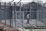 A man, whose faces is obsucred, walks around a concrete area surrounded by high metal fenced walls.