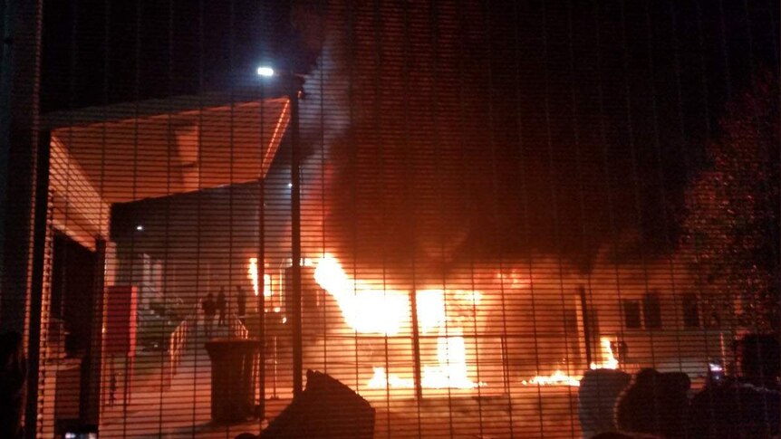 Fire consumes a building with three people standing close-by on a footpath.