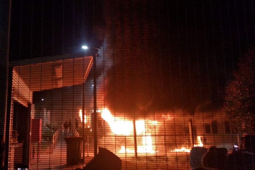 Fire consumes a building with three people standing close-by on a footpath.