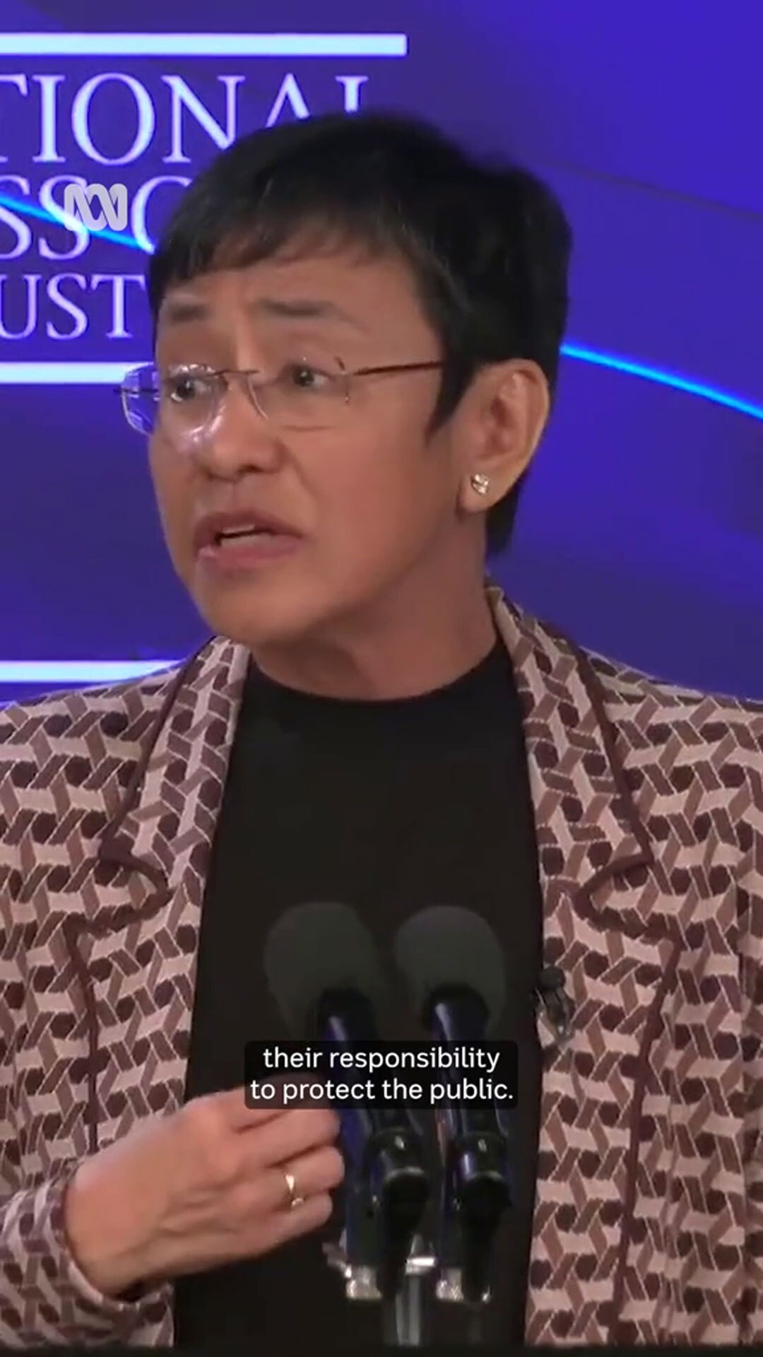 Maria Ress in a patterned coat and black top stands before two mic in front of a branded backdrop