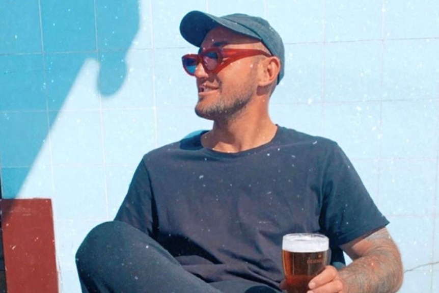 A young man wearing a cap and red sunglasses smiles while sitting in a chair, holding a beer.