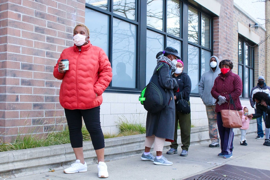 A woman in a red coat holding a coffee cup stands in line outside a building