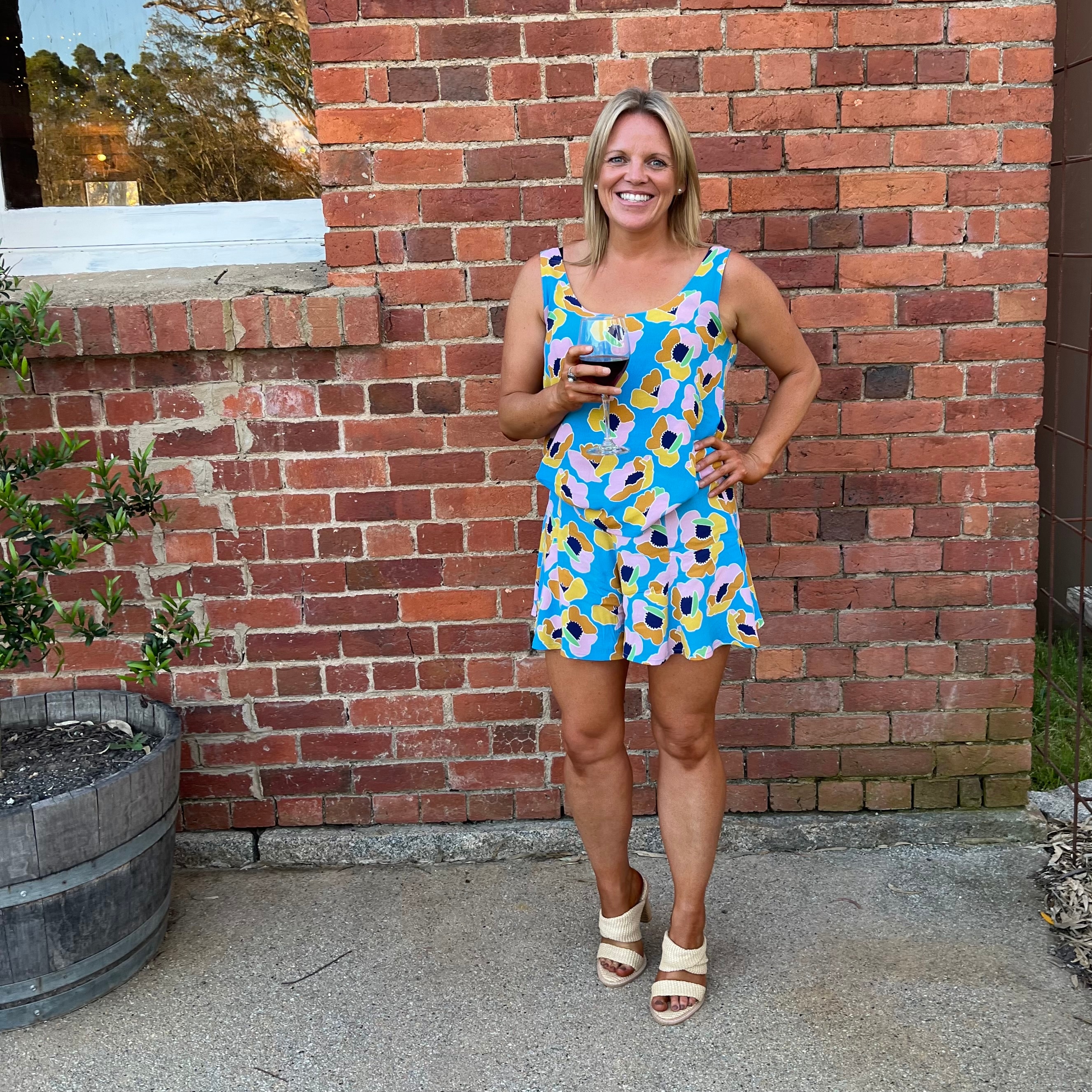 A confident, smiling blonde woman standing in front of a brick wall holding a glass of red wine.