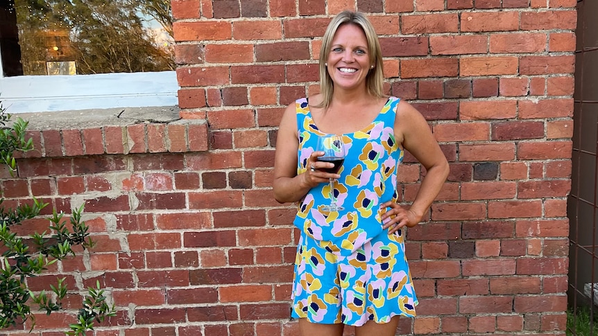 A confident, smiling blonde woman standing in front of a brick wall holding a glass of red wine.