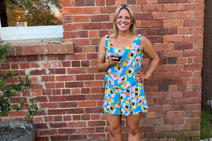 A confident, smiling blonde woman standing in front of a brick wall holding a glass of red wine.