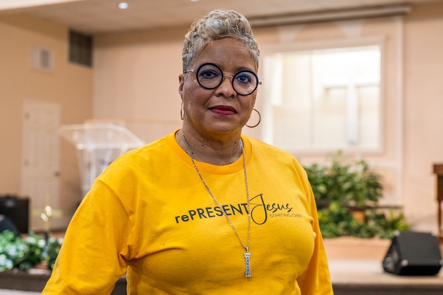 A Black woman smiles while wearing circle glasses and a yellow tshirt with rePresent Jesus written on it