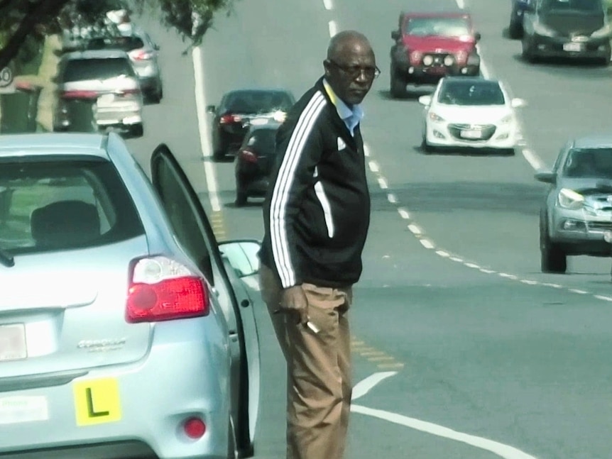 A man in a tracksuit top gets out of a car with 'L' plates on it in a busy street