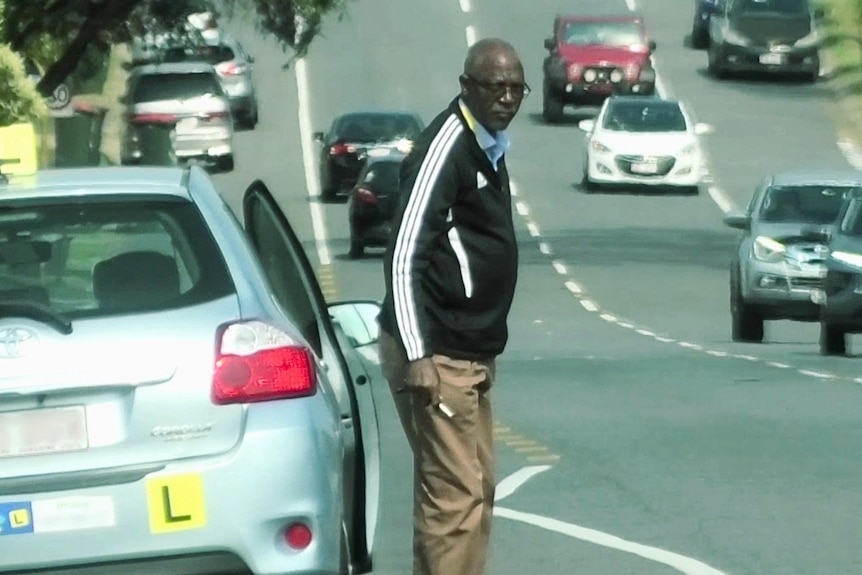 A man in a tracksuit top gets out of a car with 'L' plates on it in a busy street