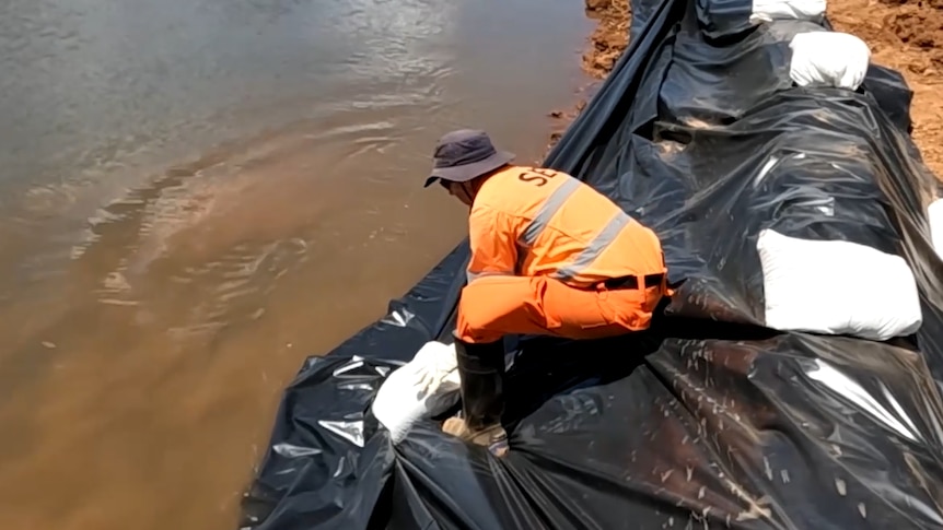 Man wearing orange ses uniform puts sand bags on black tarp to seal a levee against floodwaters