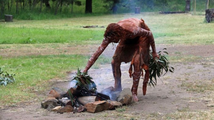 An Indigenous Australian in traditional dress bends over to put leaves on a fire