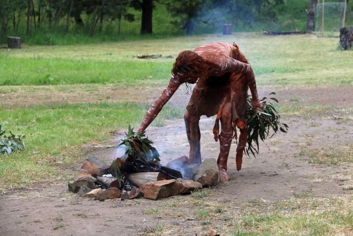 An Indigenous Australian in traditional dress bends over to put leaves on a fire