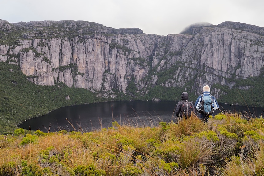 Two hikers walk towards a lake in Tasmania's Tyndall Ranges , with a large stony cliff backdrop.