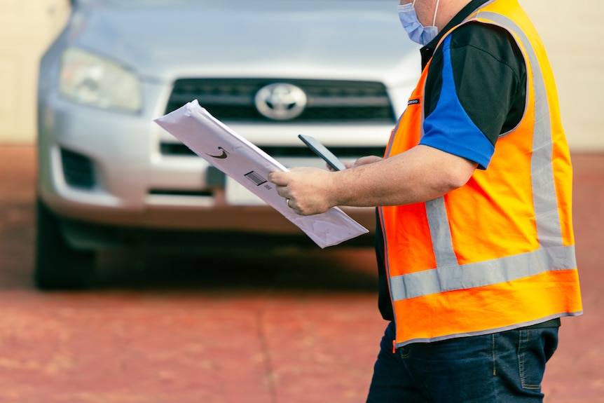 A man in an orange high-vis vest carries an envelope as he walks past a car on a suburban street. 