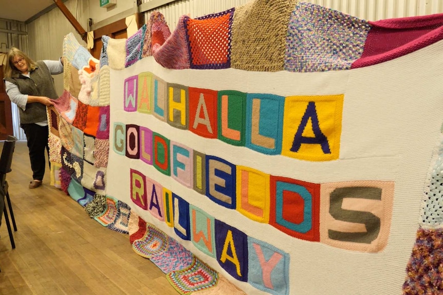 A large colourful blanket that says Walhalla Goldfields Railway, being held up by women.