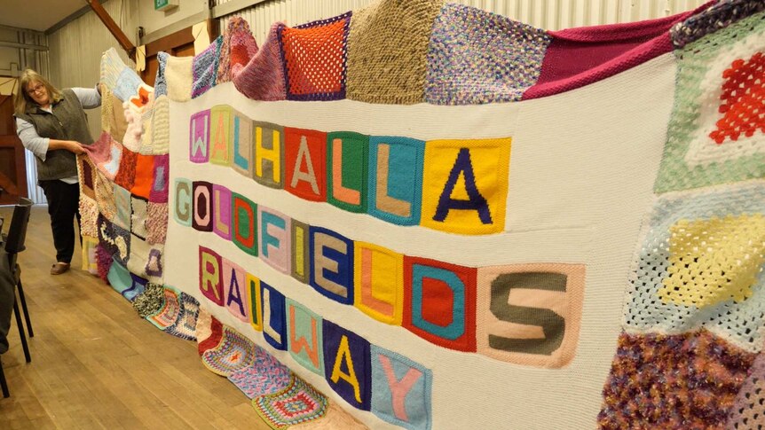 A large colourful blanket that says Walhalla Goldfields Railway, being held up by women.