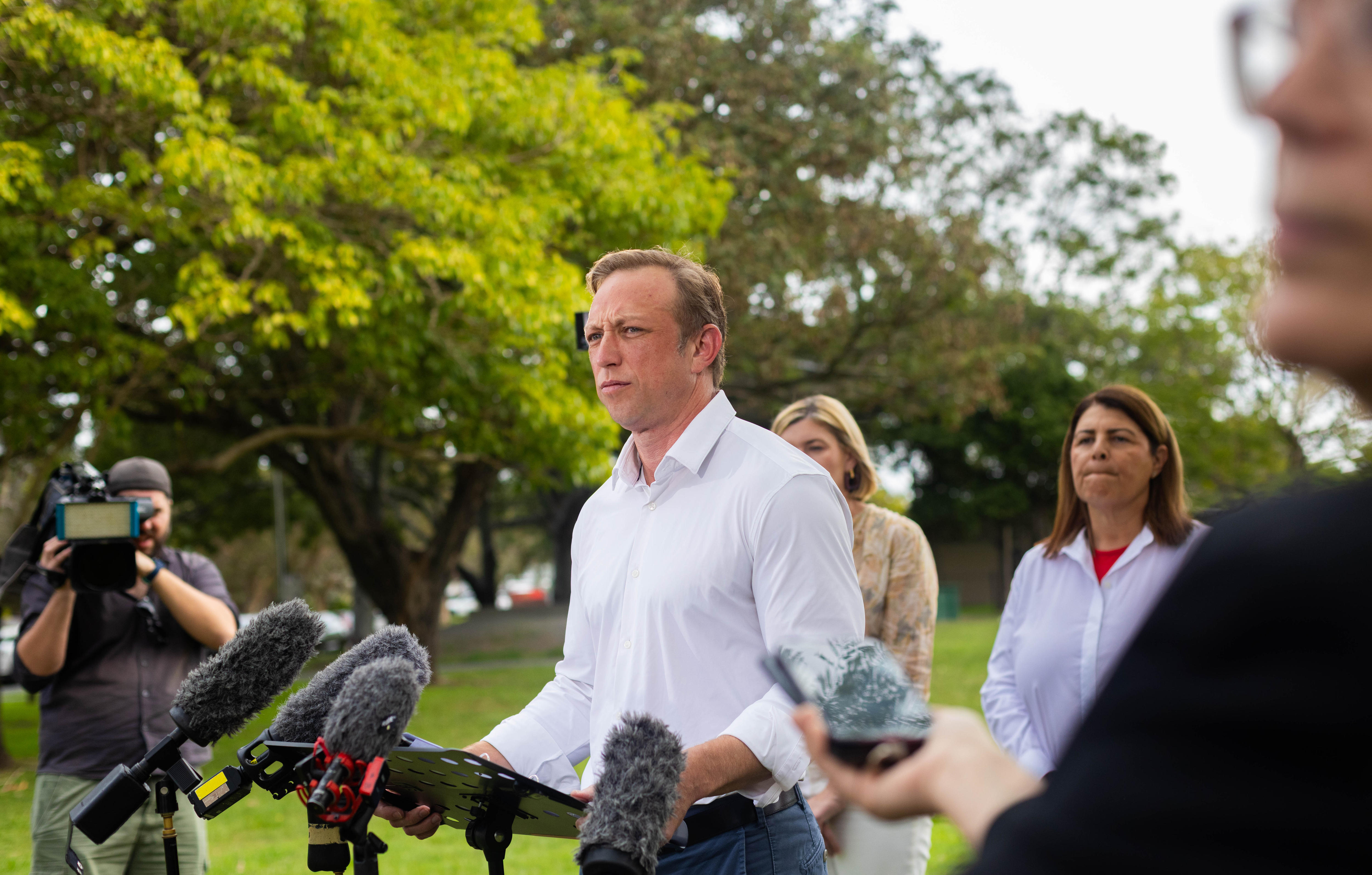 A man in a white buttoned shirt standing in front of press microphones with trees in the background.