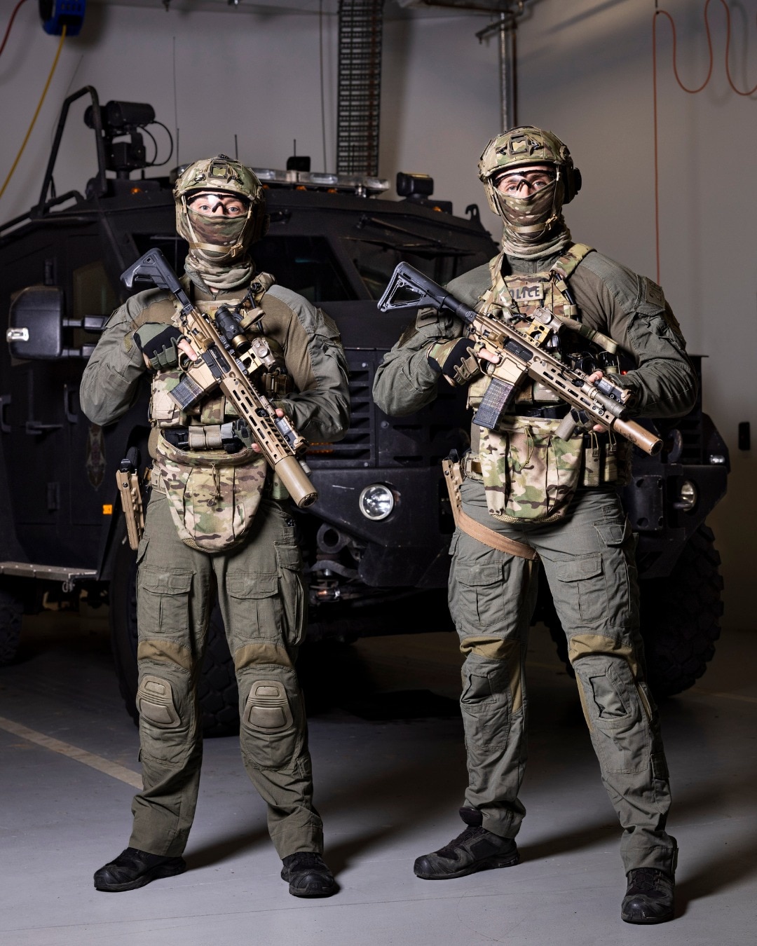 Two men in camouflaged armour hold automatic rifles and stand in front of an armoured vehicle.