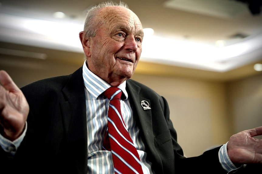 An elderly man wearing a striped blue shirt, striped red tie and black jacket gestures with his hands in a conference room.