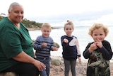 Indigenous woman sitting on rock with three young children holding shells at a rocky beach, headland in background
